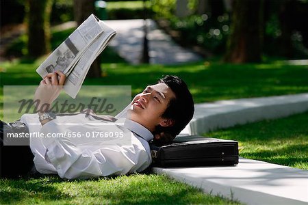 A man lies down and reads the newspaper in the park