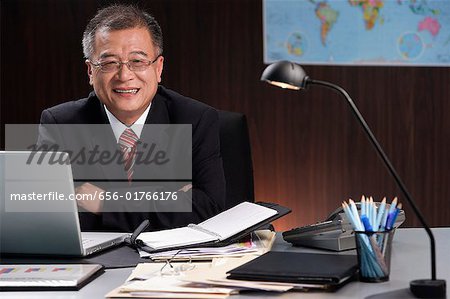 Un homme sourit à la caméra, car il est assis à son bureau