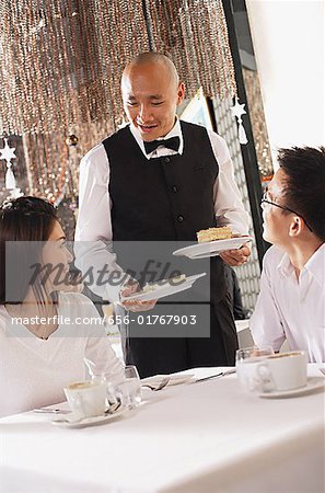 Couple in restaurant, waiter standing next to their table, holding plates of dessert