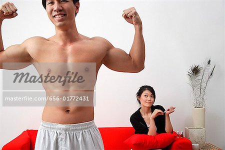 Couple à la maison, l'homme au premier plan flexing muscles, femme assise sur le canapé