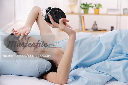 Young woman lying on bed, frowning at alarm clock