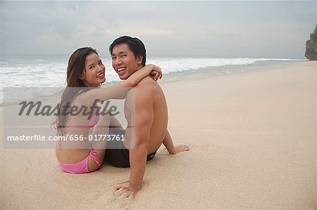 Couple sitting on beach, embracing, looking at camera