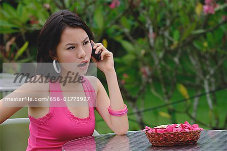 Young woman sitting at table outdoors, on the phone, angry expression on face