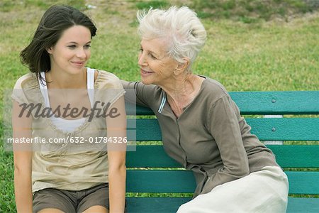 Grandmother and teenage granddaughter sitting together on bench, both smiling