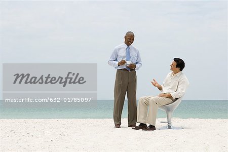 Two business associates chatting on beach, one holding coffee cup