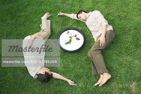 Two men lying on grass around bowl with flower, eyes closed