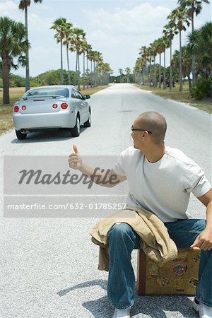 Male hitchhiker sitting on suitcase on road, looking over shoulder at passing car