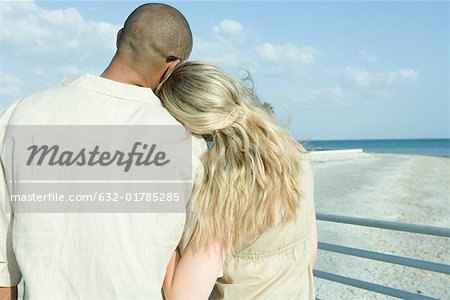 Couple standing arm in arm looking at view, woman's head on man's shoulder, rear view