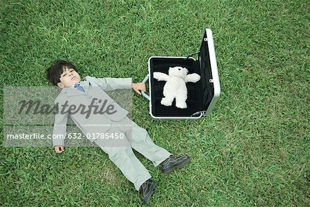 Little boy in full suit, lying on grass, next to open briefcase containing teddy bear