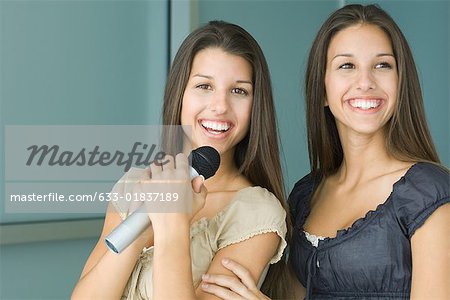 Teenage twin sisters side by side, both smiling, one holding microphone