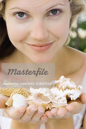 Woman holding up handfuls of seashells, smiling at camera