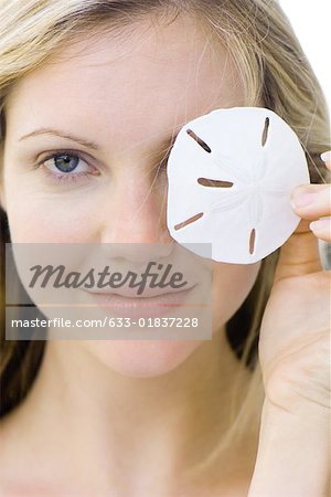 Woman covering one eye with sand dollar, smiling at camera, portrait