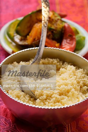 Couscous in silver bowl, plate of vegetables behind