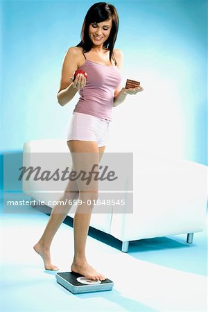 Young woman standing on scales with piece of cake and apple