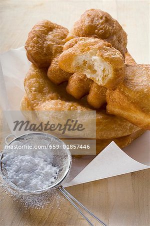 Fried pastries on paper, icing sugar in sieve