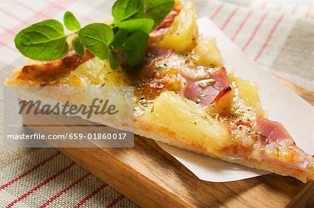 Slice of Hawaiian pizza with fresh oregano on chopping board