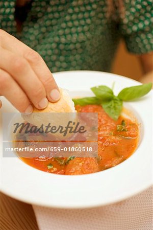 Hand dipping white bread into tomato soup with basil