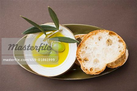 Green olives on twig in bowl of olive oil, white bread