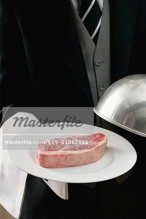 Butler serving raw beef steak on plate with dome cover