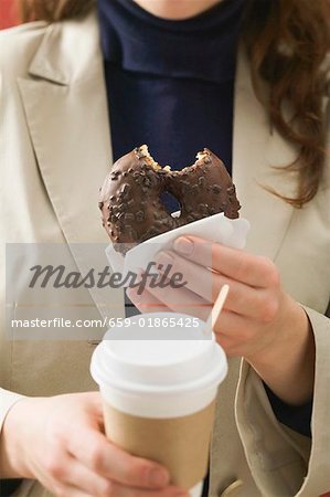 Woman holding chocolate doughnut with bite taken & cup of coffee