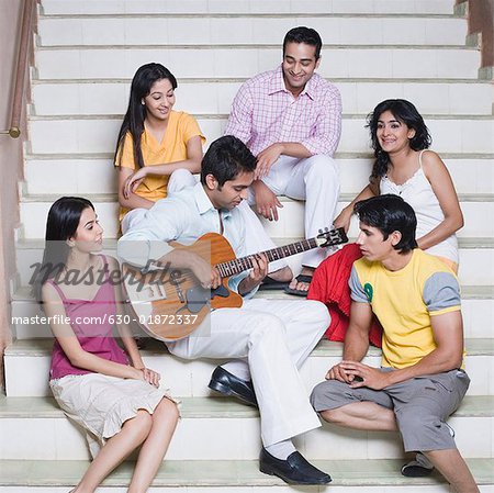 Junger Mann mit seinen Freunden sitzt neben ihm eine Gitarre spielen