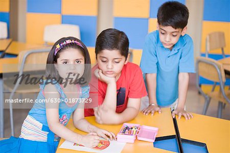 Portrait of a girl drawing on a sheet of paper with two boys looking at her drawing sheet