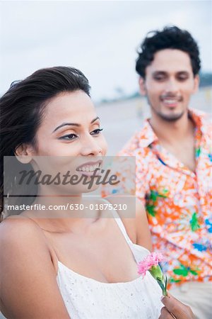 Close-up of a young woman holding a flower and smiling with a young man standing behind her