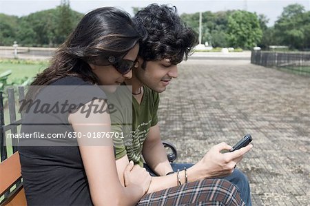 Side profile of a young couple sitting on a bench and operating a mobile phone, Goa, India