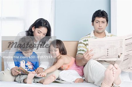 Young woman teaching her children with her husband sitting beside them and reading a newspaper