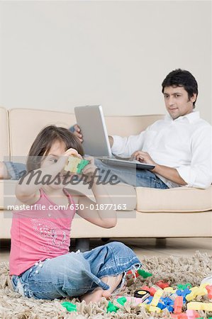 Girl playing with toys with her father sitting behind her and looking at her