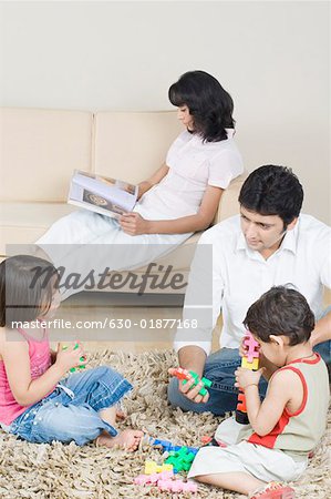 Mid adult man playing with his children and his wife reading a magazine in the background
