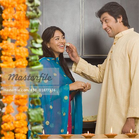 Side profile of a mid adult man feeding a piece of burfi to a young woman in a domestic kitchen and smiling