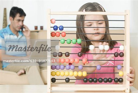 Girl playing with an abacus with her father talking on a mobile phone in the background