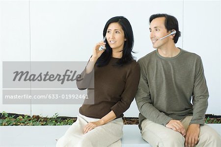 Paar sitzen nebeneinander, Frau mit Handy, Mann mit Headset, beide Lächeln