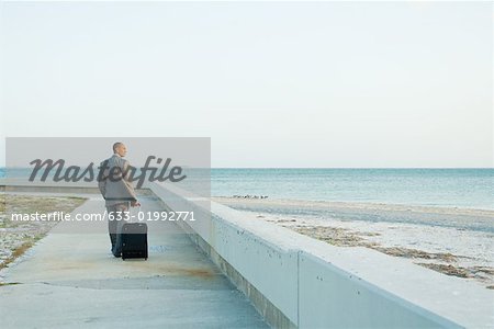 Homme d'affaires marchant sur le trottoir à la plage, en tirant la valise derrière lui, regardant la vue