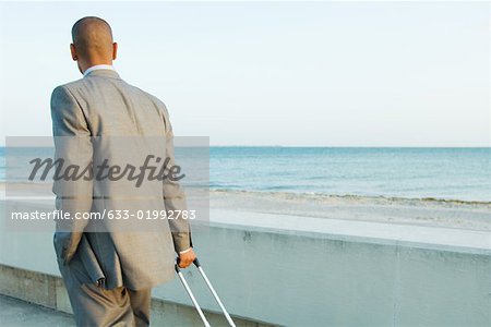 Businessman walking by the sea, pulling suitcase, looking at view