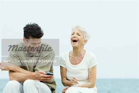 Femme senior et fils adulte assis côte à côte à l'extérieur, en riant, man holding cell phone