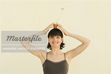 In die Kamera lächelnde Frau hält kleine Topfpflanze auf Kopf, Porträt