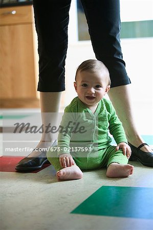 A baby sitting on kitchen floor with his mom behind him.