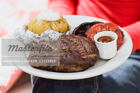 Woman holding plate of steak, baked potato, vegetables & sauce