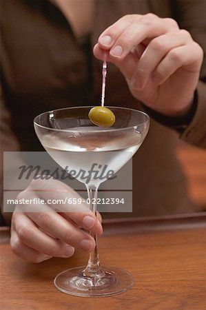 Woman holding olive on cocktail stick over glass of Martini