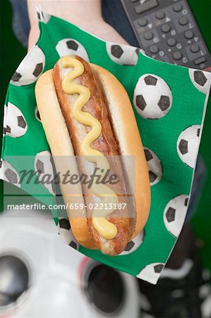 Hand holding hot dog with mustard on napkin with football motifs