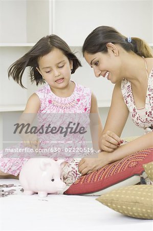 Girl putting a coin into a piggy bank with her mother sitting beside her and smiling