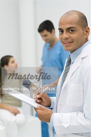Doctor writing on clipboard, smiling at camera, nurse measuring patient's blood pressure in background