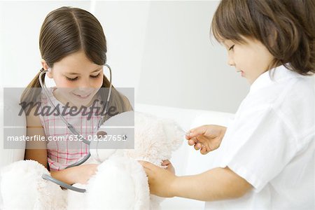 Two children playing doctor, girl listening to teddy bear's heart with stethoscope