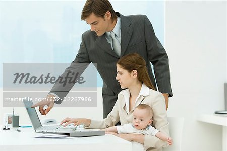 Professional woman in office, holding baby on lap, looking at laptop with male colleague