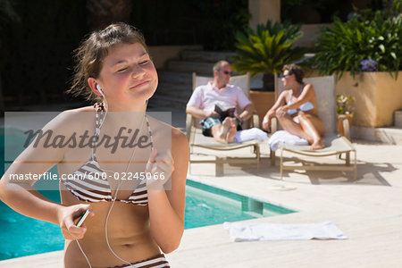 Teenaged Girl Sitting by Swimming Pool, Listening to Music, Parents in the Background Teenaged Girl Sitting by Swimming Pool, Listening to Music, Parents in the Background