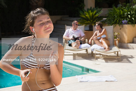 Teenaged Girl Sitting by Swimming Pool, Listening to Music, Parents in the Background