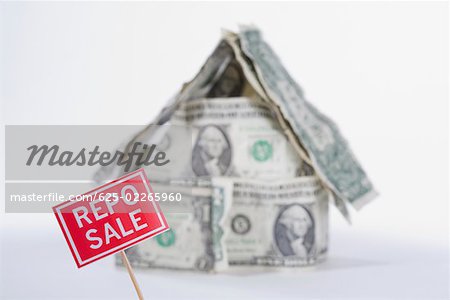 For Sale board in front of a miniature house made up of US dollar bills