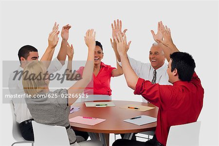 Three businessmen and two businesswomen smiling with their arms raised in a meeting